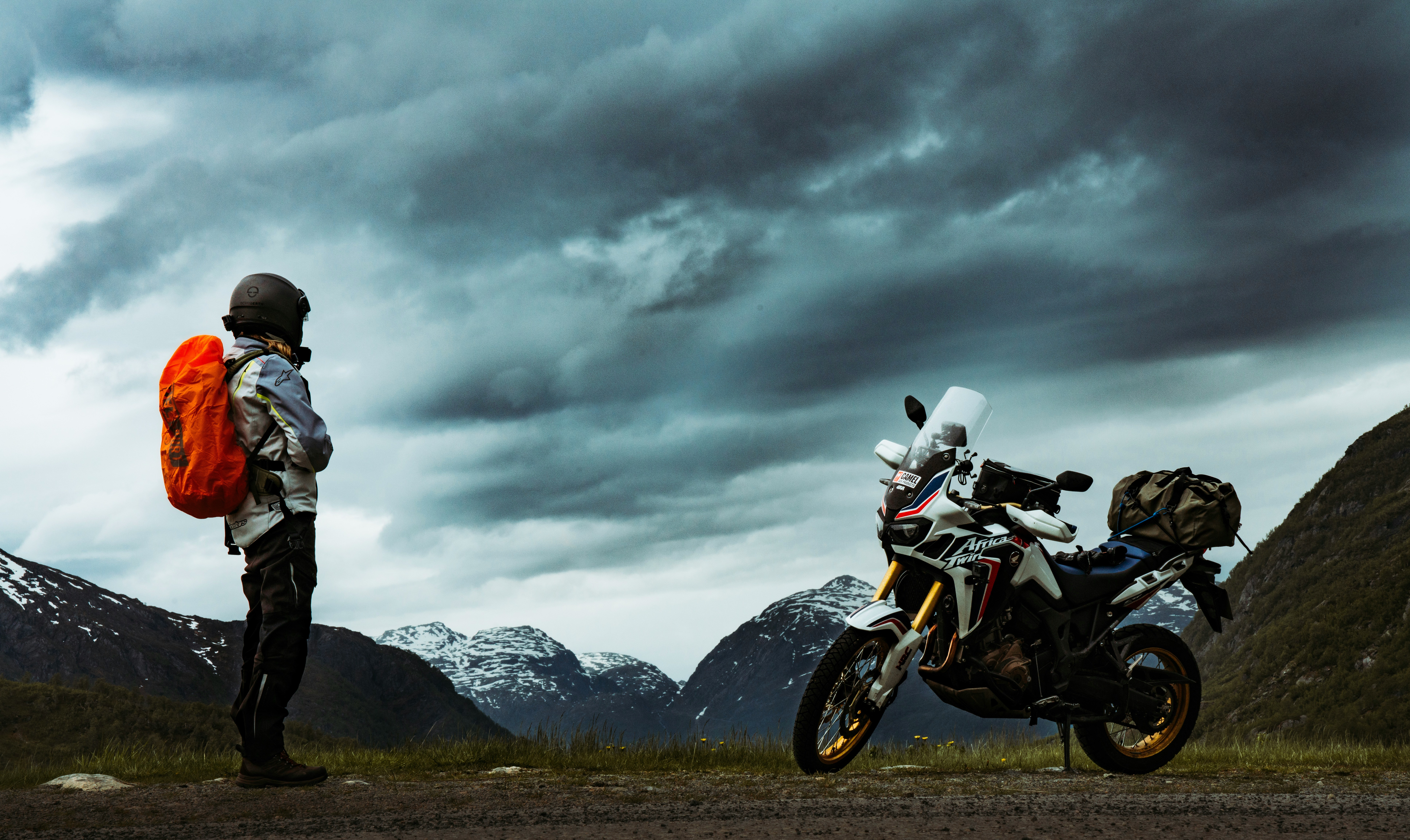 Rider beside a loaded adventure motorcycle in a mountain valley under dark clouds